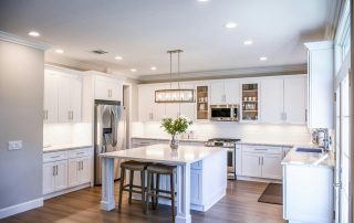 Esta imagen muestra una cocina espaciosa y bien iluminada con un diseño minimalista. Destacan los gabinetes blancos, una isla central con taburetes, y electrodomésticos de acero inoxidable, complementados por encimeras de mármol y una iluminación sutil. Perfecta ilustración de cómo combinar funcionalidad y estilo en el diseño de cocinas modernas.